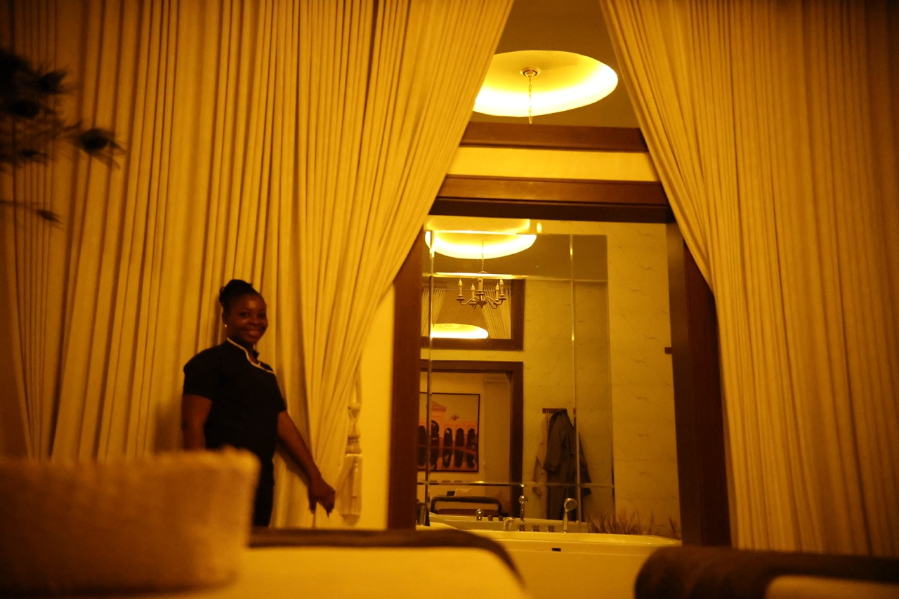 A GoldCat staff member smiling at the threshold of a treatment room, curtains framing the doorway, jacuzzi visible in the background, candlelit warm interior