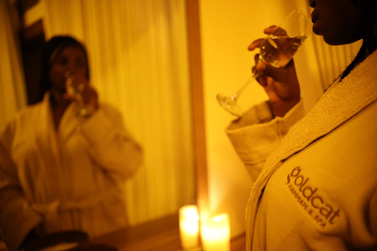 A solo woman in a GoldCat-branded white robe holding a glass, candlelit warm interior with mirror reflection