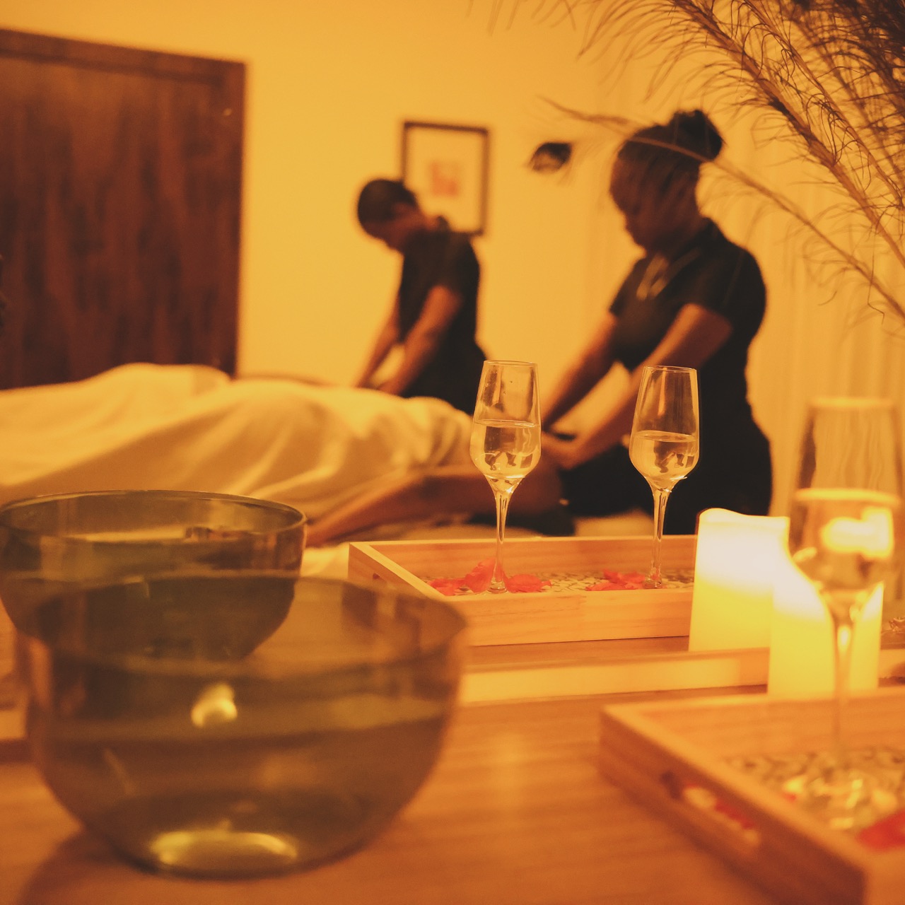 A couples body treatment in the GoldCat treatment room: two therapists working in soft candlelight, glass bowls of water and small champagne flutes set out in the foreground