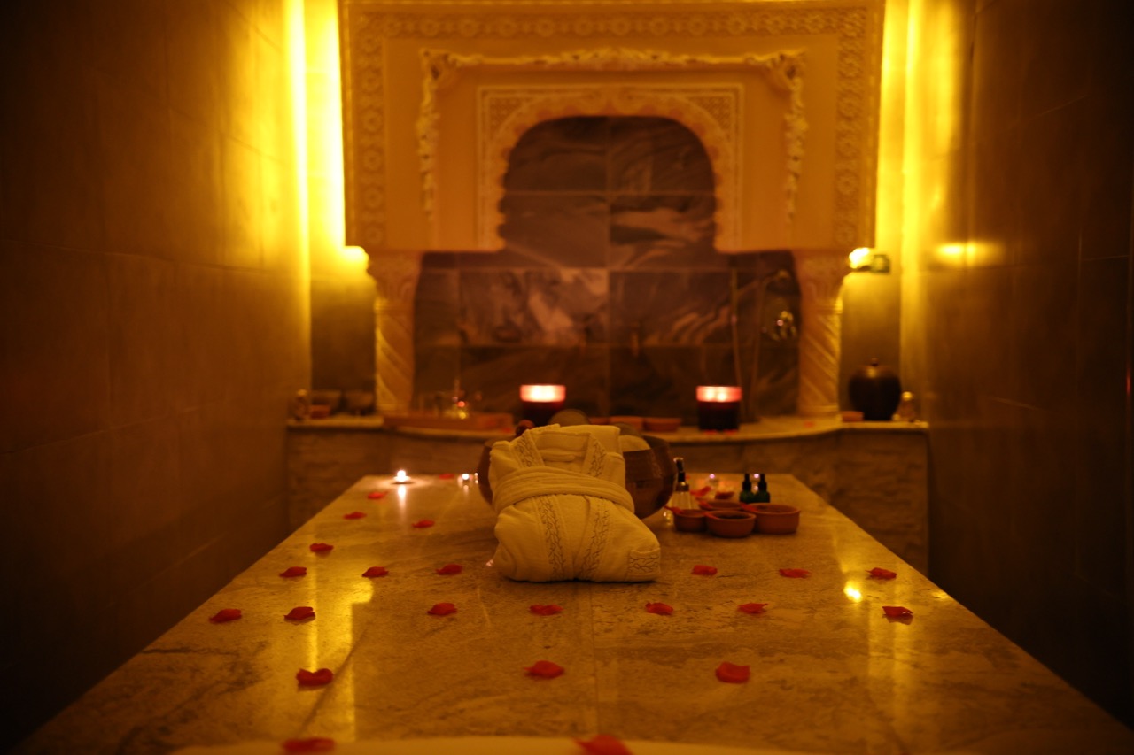 The marble hammam room at GoldCat: warm marble slab, scattered rose petals, ornate Moroccan stucco niche, candlelight and terracotta tea bowls