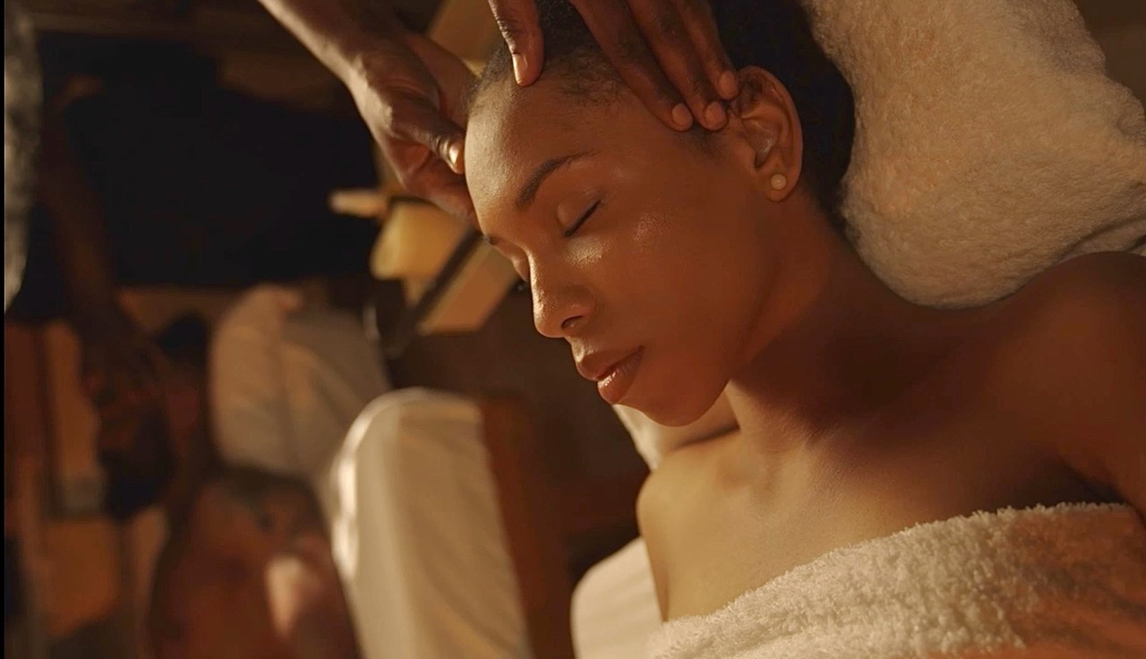 A guest receiving a facial: hair wrapped in a white towel, eyes closed, therapist's hands gently working at the temples